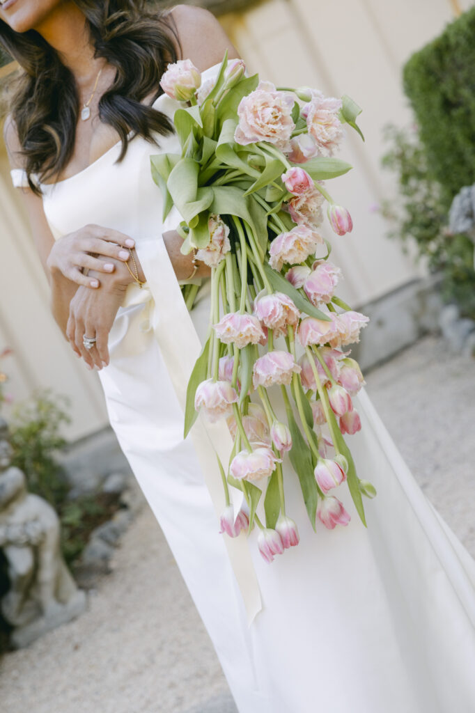 Close-up of a bride holding a cascading bouquet of soft pink tulips against her white off-the-shoulder wedding gown at Stonepine Estate in Carmel Valley