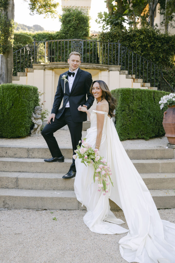 Bride and groom posing playfully on the stone garden steps at Chateau Noel at Stonepine Estate, surrounded by sculpted hedges, garden statuary, and a wrought iron balcony, the bride holding a cascading pink tulip bouquet
