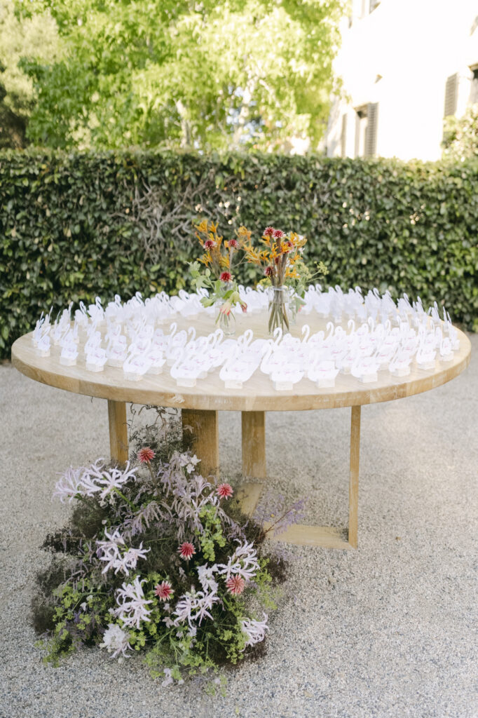 Circular wood table displaying white swan-shaped escort cards arranged in rows with a wildflower centerpiece, set on the gravel garden terrace at Stonepine Estate with a manicured hedge backdrop and low floral ground arrangement beneath the table