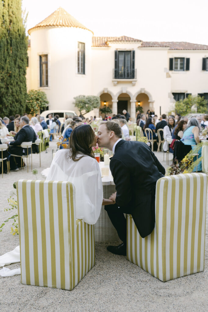 Bride and groom seated together at their sweetheart table in yellow and white striped high-back chairs in the courtyard of Chateau Noel at Stonepine Estate, guests and the villa visible behind them at dusk