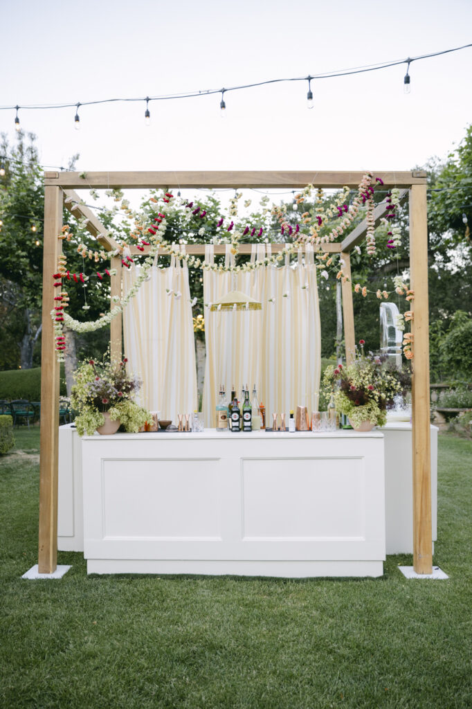 Outdoor wedding bar at Stonepine Estate with a wood pergola frame draped in cream ribbons and colorful floral garlands, set on the lawn at dusk with string lights overhead and garden greenery in the background