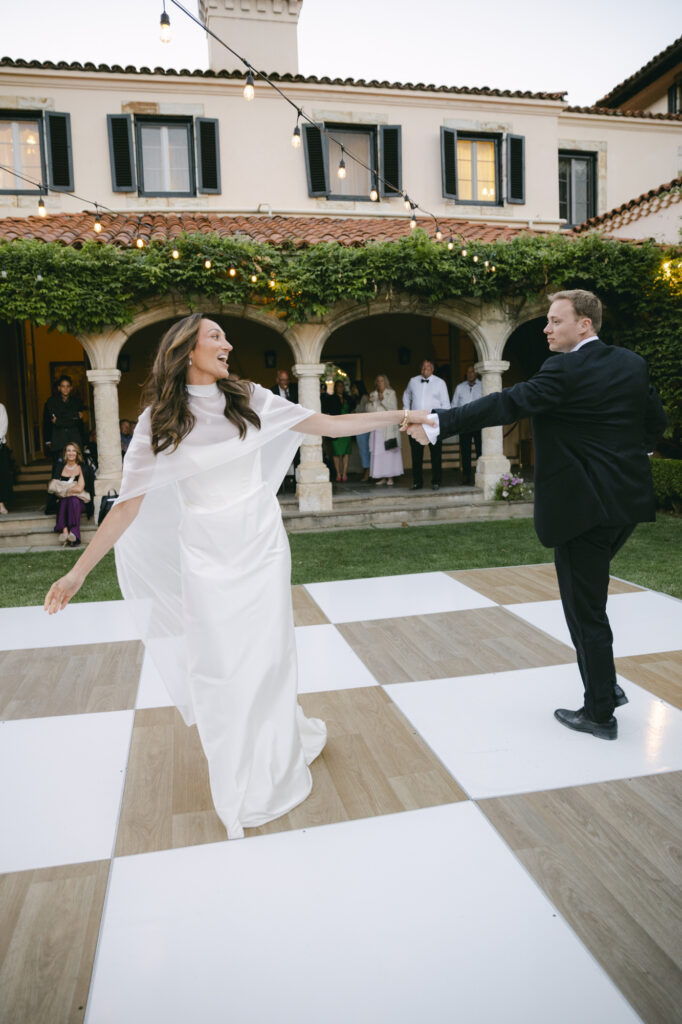 Bride and groom sharing their first dance on a checkered black and white dance floor in the courtyard of Chateau Noel at Stonepine Estate, string lights and ivy-draped arches glowing behind them at dusk