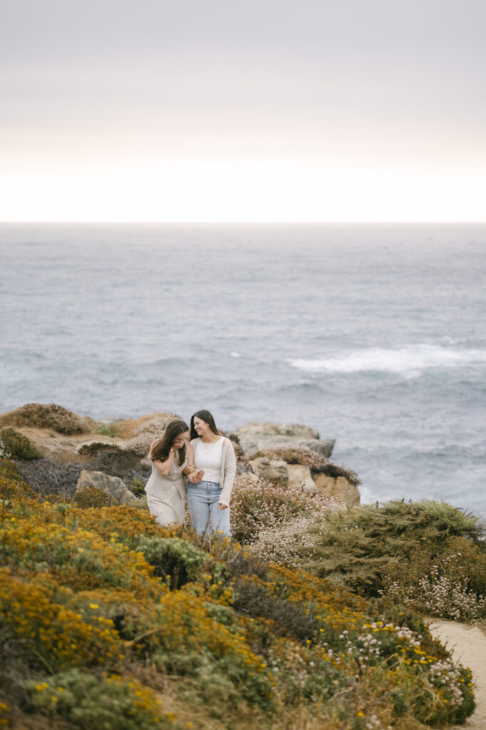 Wide shot of engaged couple among wildflowers, admiring their engagement ring, and coastal rocks at Garrapata State Park in Carmel