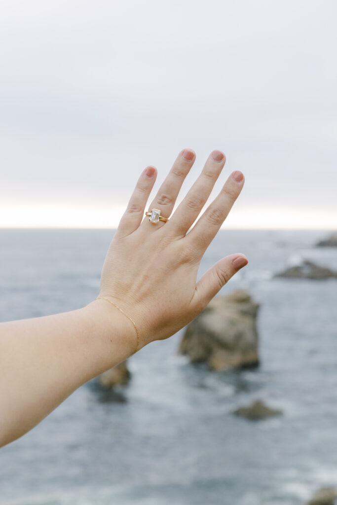 Engagement ring on hand raised against ocean backdrop at Garrapata State Park near Carmel