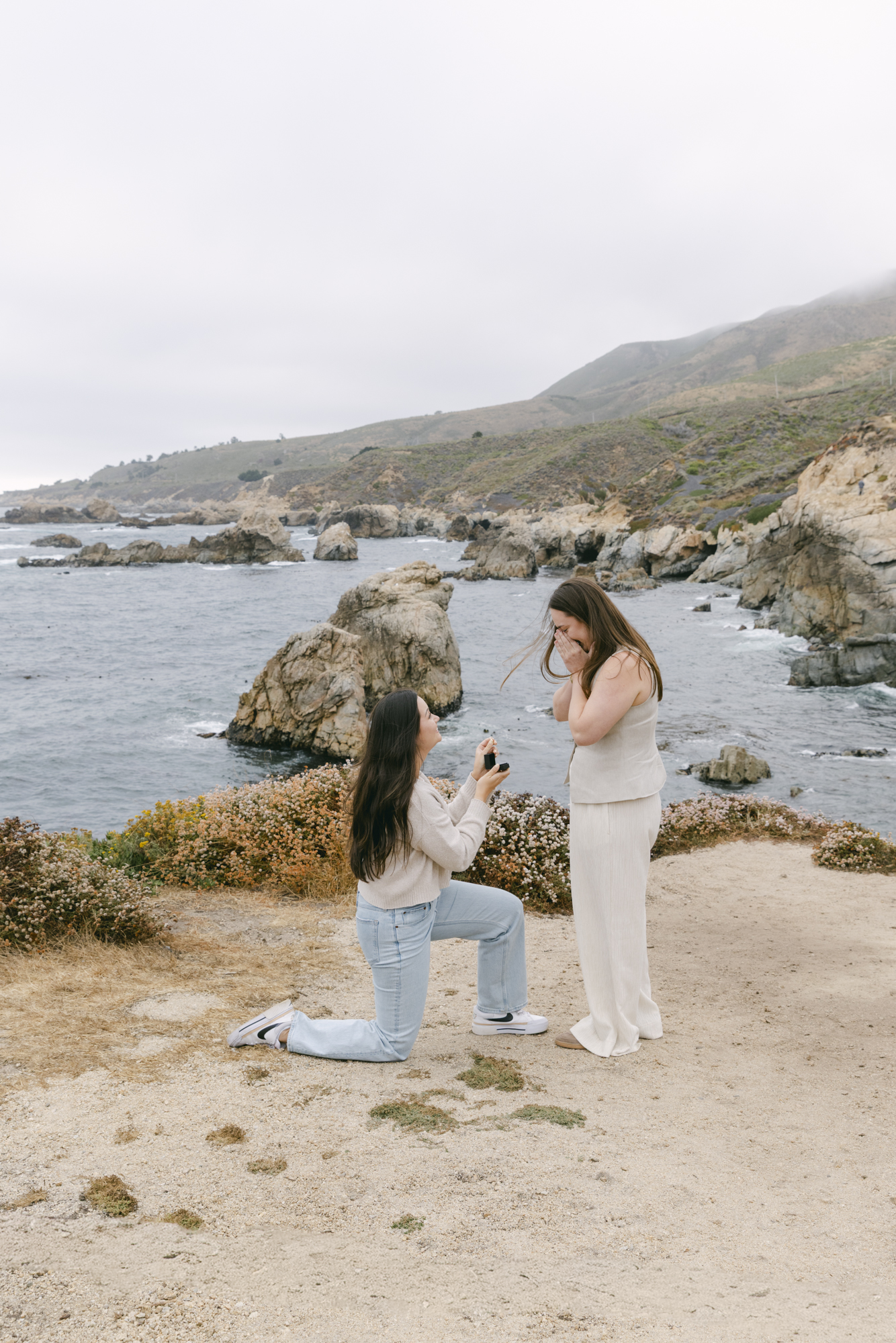 Woman kneeling to propose with ring box on rocky coastline at Garrapata State Park, her partner covering her face in surprise