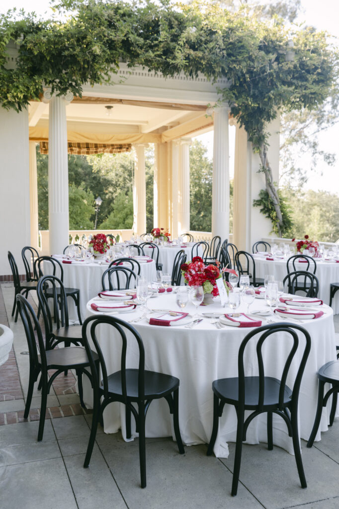 Round reception table with black bentwood chairs and red floral centerpieces set beneath Villa Montalvo's columned pergola at a luxury wedding