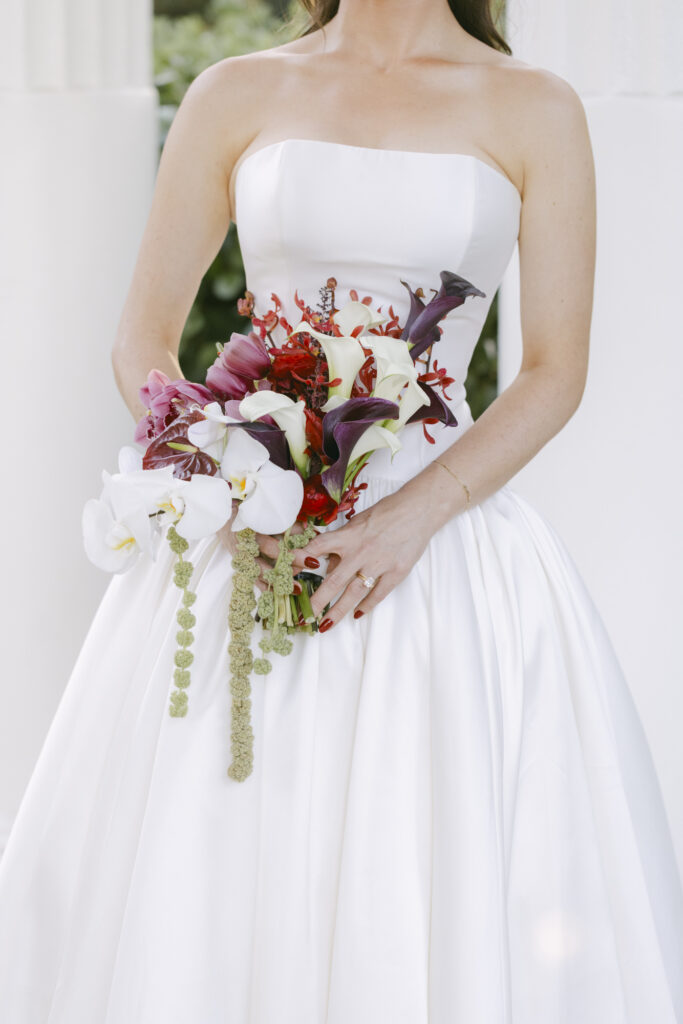 Close up of a bridal bouquet with white orchids, calla lilies, and deep red and burgundy florals against a strapless white wedding gown